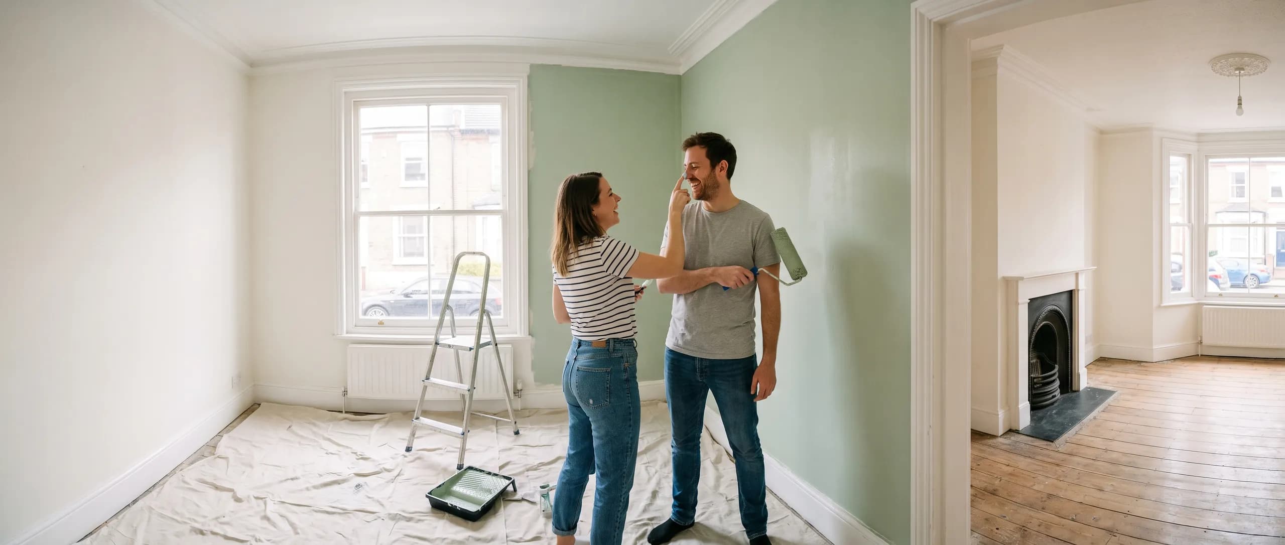 A couple laughing as they paint the living room of their new flat sage green, dust sheets and a stepladder on the floorboards.